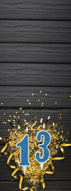 Number 13 blue celebration candle and gold confetti on dark wooden background. 13th birthday card. Anniversary and birthday concept. Vertical banner. Copy space