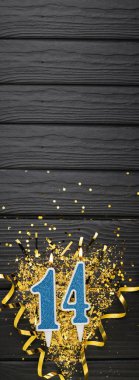 Number 14 blue celebration candle and gold confetti on dark wooden background. 14th birthday card. Anniversary and birthday concept. Vertical banner. Copy space