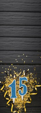 Number 15 blue celebration candle and gold confetti on dark wooden background. 15th birthday card. Anniversary and birthday concept. Vertical banner. Copy space