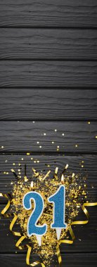 Number 21 blue celebration candle and gold confetti on dark wooden background. 21th birthday card. Anniversary and birthday concept. Vertical banner. Copy space