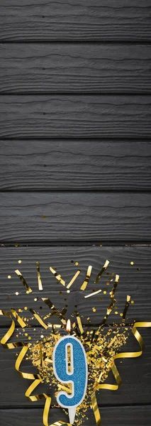 Number 9 blue celebration candle and gold confetti on dark wooden background. 9th birthday card. Anniversary and birthday concept. Vertical banner. Copy space