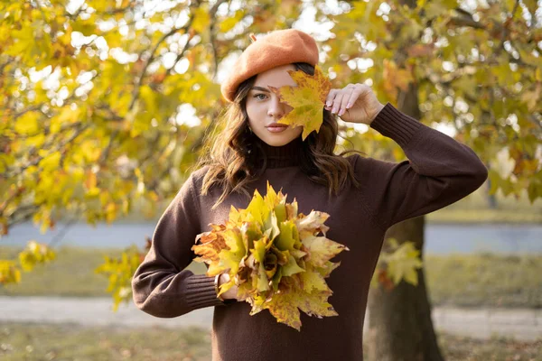 stock image Portrait of a beautiful autumn woman standing near colorful autumn leaves. Pretty happy girl looking at camera holding in her hands yellow maple leaves covering her eye. Copy space