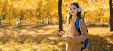 Sonbahar parkında sırt çantalı kız öğrenci kulaklıkla müzik dinliyor ve akıllı telefonunu elektrik bankasıyla şarj ediyor. Pankart. Boşluğu kopyala