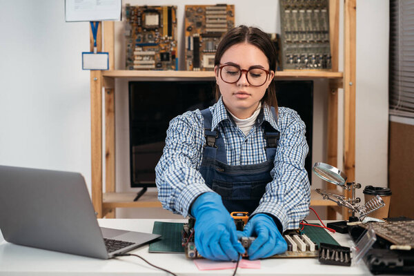 Tech fixes motherboard in service center. Female repairman working with computer with a part in hands. Monitors and other laptops in the background waiting for service. Copy space