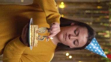 Güzel, mutlu, genç bir kadın elinde doğum günü pastası tutan 65 numaralı süveter giyiyor. Çakmakla yanan altın mumlar. Doğum günü ve yıldönümü kutlaması kavramı. Bakış açısı. Dikey video