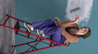 Mutsuz, üzgün, yorgun kadın evdeki duvarı boyuyor, merdivenlerde dinleniyor. Dikey video. Boşluğu kopyala