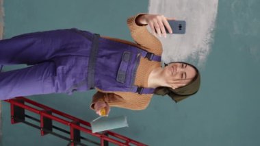 Cep telefonu ve boya silindirli boyacı kadın, mavi ev duvarında çalışıyor. Ev dekorasyonu ve iç yenileme konsepti. Dikey video