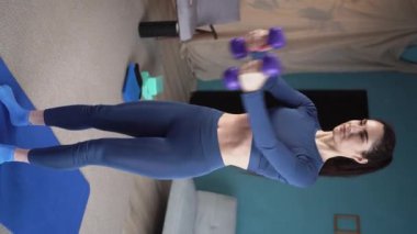 Spor kıyafetli güzel bir kadın evde halterli egzersiz yapıyor. Kadınlar halterleriyle evde kilo kaybediyor. Spor ve eğlence konsepti. Dikey video