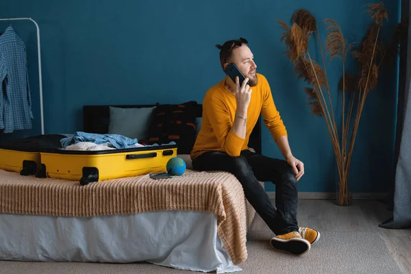 Attractive traveler man with suitcase sit on cozy comfort bed inside modern apartment room speak on cellular, making hotel reservation on phone from home, copy space