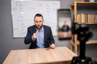 Erkek öğretmen çevrimiçi İngilizce dersi veriyor, akıllı telefon kullanıyor, ana ofiste gramer kurallarıyla kara tahtanın önünde oturan webcam 'i işaret ediyor. Uzaktan eğitim konsepti. Boşluğu kopyala