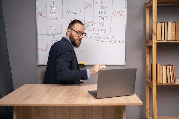 stock image Serious teacher man with marker at whiteboard watch on laptop webcam in classroom interior at home. Online lesson of English language, repetition of rules. Copy space