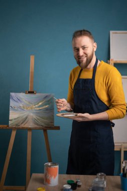 Sanat atölyesinde çalışan kaygısız erkek ressam. Resminin yanında dururken kameraya gülümseyen mutlu yetişkin sanatçı. Yaratıcı adam tuvalde yeni bir resim yapıyor. Boşluğu kopyala