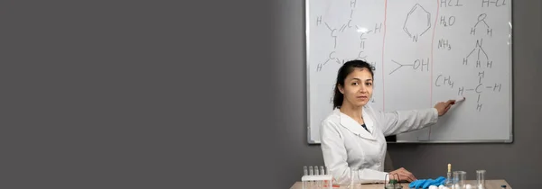 A Chemistry teacher sitting at desk and pointing on blackboard, teaching in a classroom setting. Banner. Copy space
