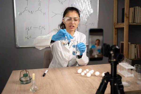 Online teacher, mentor. School science classroom. Teacher explains chemistry. Young woman teaches and records video lessons, smartphone on a tripod in front of her. Copy space