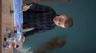 Öğrenci bir çocuk evde bir masada oturuyor ve tuvale akrilik boyalarla resim yapıyor. İlham, eğitim ve sanat konsepti. dikey video. Boşluğu kopyala.