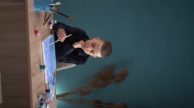 Öğrenci bir çocuk evde bir masada oturuyor ve tuvale akrilik boyalarla resim yapıyor. dikey video. Boşluğu kopyala.