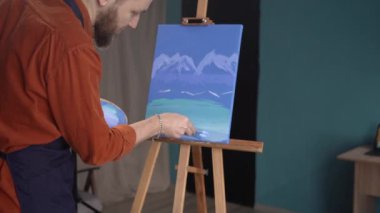 Yakın plan, beyaz sakallı, önlüklü bir adam ev stüdyosunda, dikiz aynasındaki tuvale resim çiziyor. Boş zaman, yaratıcı hobi ve sanat konsepti. Boşluğu kopyala.