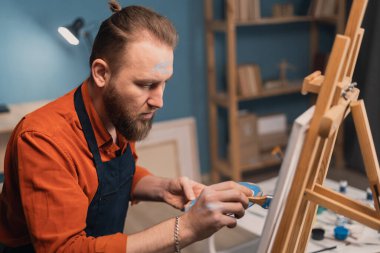 Kafkas sakallı, önlüklü bir ressam sandalyede oturur ve yağlı fırçalı bir resim çizer. Tablo bir sehpanın üzerine kurulmuş. hobi ve tatil konsepti.