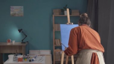 Bir sehpanın yakınındaki bir odada duran genç ve mutlu bir kadının arka görünüşü elinde boya ve fırçalarla bir palet tutuyor ve soyut bir resim çiziyor. Boşluğu kopyala.