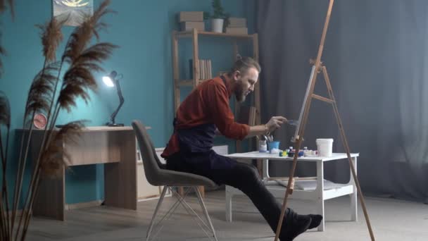 大胡子千禧年艺术家在围裙上工作的肖像 坐在抽象画上 用画笔创作现代侧视图画 在创意工作室的画架上画布 垂直视频 — 图库视频影像