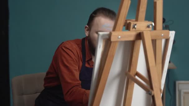 一个在围裙上工作的大胡子千禧年艺术家的肖像 坐在抽象画上 用画笔创作现代侧视图画 在创意工作室的画架上画布 垂直视频 — 图库视频影像