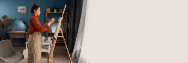 Stüdyodaki genç Brezilyalı bir kadın ressam tuvalin yanında duruyor ve fırçalı bir tabloya boya sürüyor. Siyah saçlı, günlük kıyafetler giymiş. Yaratıcılık ve hobi kavramı. Pankart. Boşluğu kopyala.