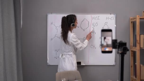 教師ブロガーは スマートフォンでライブ教育ビデオを記録します マーカーボードに立つと化学式が書かれ意味がわかります 化学の授業をオンラインで ブログのコンセプト — ストック動画