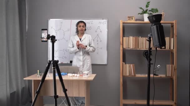 スマートフォンは学校でビデオレッスンを記録します イタリアの若い化学教師が教室の式を説明する白いコートの黒板に立っています オンライン学習 — ストック動画