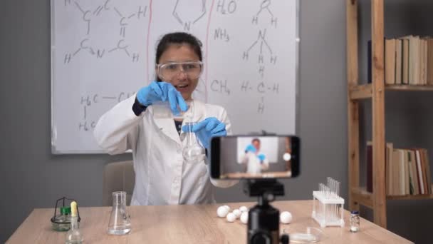 远程远程电子学习 化学老师在网上授课 戴着橡胶手套 进行化学搅拌两种液体 在智能手机上记录一个教训 — 图库视频影像