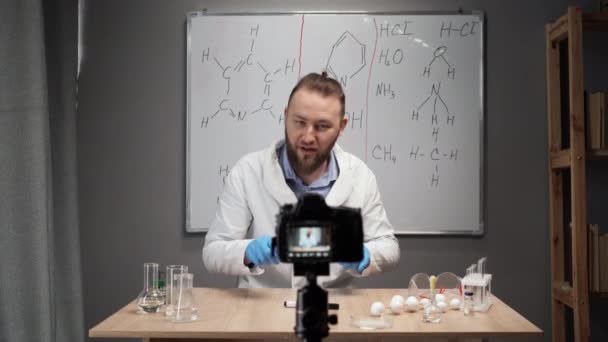 在线导师化学老师记录Vlog在家里的家庭视频工作室 一个留着胡子的白衣男子坐在一块白板的后面 指黑板上关于公式的谈话 — 图库视频影像