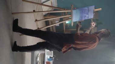 Önlük giymiş yetenekli beyaz bir sanatçı enerjik ve hevesli bir şekilde fırçayla modern bir sanat eseri yaratıyor. Evde yaratıcı stüdyo. En sevdiğim hobi. dikey video. Boşluğu kopyala