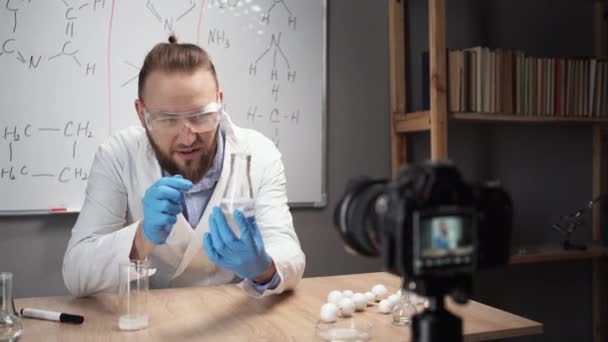 在线导师化学老师记录Vlog在家里的家庭视频工作室 一个留着胡子的白衣男子坐在一块白板的后面 拿着一个瓶子讲述了一种化学反应 — 图库视频影像