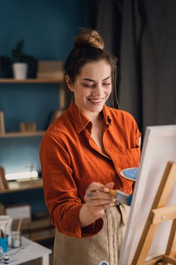 Yaratıcı çağdaş Latin ressam ev stüdyosunda sehpanın yanında dururken renkli ve soyut tablolar çiziyor. Pozitif kadın sanatçı, yakın plan portre.