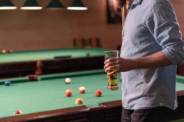 Unrecognizable man with a glass of beer standing near the pool table with colorful billiard balls. Copy space