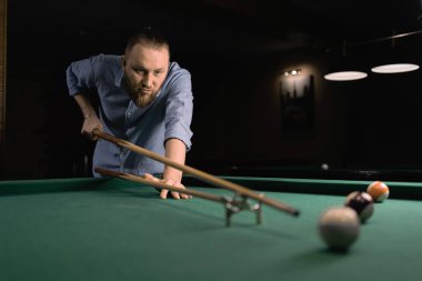 Genç sakallı profesyonel bir adam karanlık bir bilardo kulübünde bilardo oynuyor. Boşluğu kopyala.