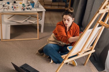 Ev sanatları atölyesinde dizüstü bilgisayarla oturmuş, online resim dersi alan bir kadın ressam. Üst Manzara. Boşluğu kopyala