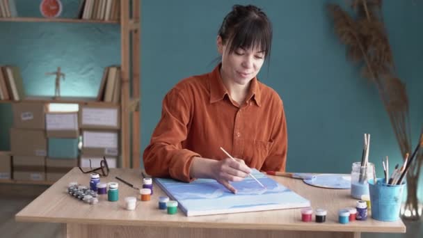 幸せな若い女性が自宅のテーブルに座っているキャンバスに絵を描き 絵具で絵を描くプロセスを楽しんでいます スペースのコピー — ストック動画