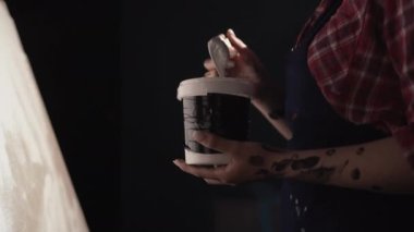 Kesilmiş kadın ressam soyut resim üzerinde çalışıyor, modern resim yaratmak için fırçayla fırçayı kovayla karıştırıyor. Büyük tuvalin önünde karanlık yaratıcı stüdyoda duran bir kadın. Yakın plan.