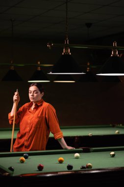 Kız oyuncu bir kulüpte bilardo oynuyor, elinde ıstakayla bilardo masasının yanında duruyor. Amerikan bilardosu. Boşluğu kopyala