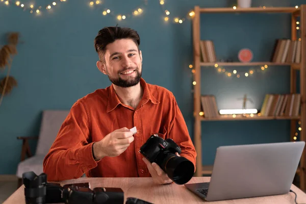 Portrait of smiling photographer holding Memory card - Flash card while sitting at the table with professional DSLR camera. Male photographer ready to insert memory card to his photo camera,copy space