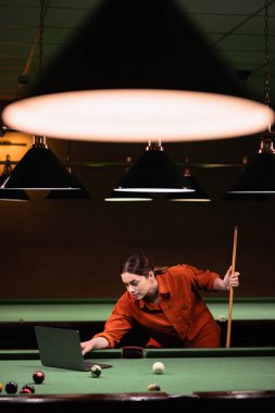 Hobiler ve boş zaman aktiviteleri. Çevrimiçi eğitim, online dersler. Genç bir kadın, karanlık bir bilardo odasında duran bilardo hakkında bir video dersi izliyor. Boşluğu kopyala