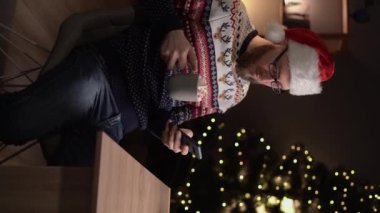 Pozitif milenyum adamı oturduğu masada cep telefonu tutacağı bardağı kullanıyor. Kapalı bir evde, Noel süsleriyle birlikte. Dikey video