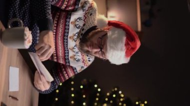 Süveter giyen ve Noel Baba şapkalı bir adam Noel mektubunu açıyor ve gece evde oturuyor. Dikey video