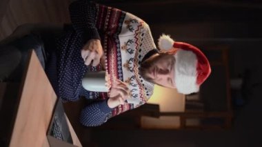 Genç adam masa başı bardağını tutarken ekranda mavi kazaklı Noel Baba şapkası giyer ve kapalı bir yerde Noel Baba 'nın evine gider. Dikey video
