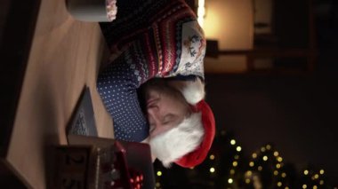 Noel Baba şapkalı genç adam geceleri çalıştığı yerde arka planda Noel ışıklarıyla otururken ellerini kaldırarak uyuyor. Dikey video