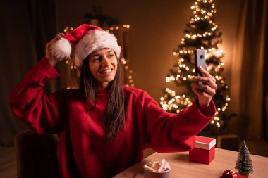 Noel kutlaması için arkadaşlarıyla ve ailesiyle birlikte akıllı telefon kullanarak evdeki oturma odasında parti yaparken gülümseyen genç kadının portresi. Blog yazarı Noel Baba. Kış tatilleri, online kutlamalar.