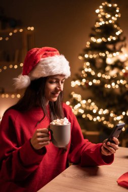 Pozitif genç bir kadının portresi evde cep telefonu tutacağı bardağı ve yılbaşı süslemeleri kullanıyor. Boşluğu kopyala