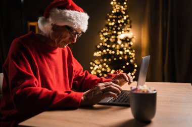 Kıdemli Noel Baba geleneksel şapkasıyla oturmuş ev ofisinin süslü odasında dizüstü bilgisayarında Noel tebriklerini yazıyor. Boşluğu kopyala
