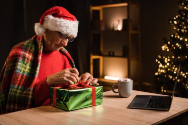 Mutlu yaşlı adam hediye paketleme masasında dizüstü bilgisayarın önünde oturuyor ve Noel arifesinde akrabalarıyla görüntülü görüşme yapıyor. Noel kutlaması kavramı