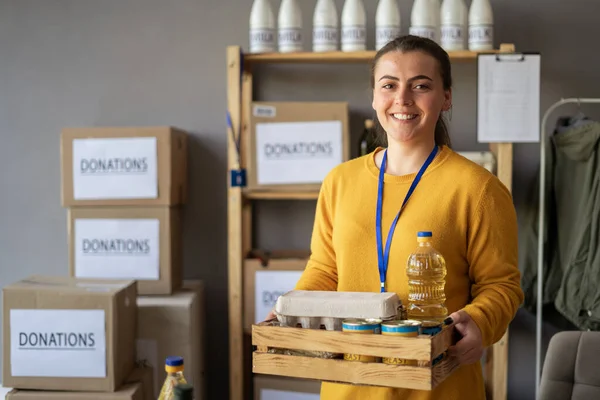 Happy smiling female volunteer holding wooden box with food at distribution or refugee assistance center. Charity, donation and volunteering concept